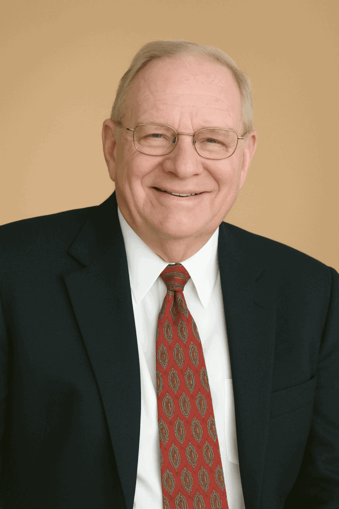 Professional portrait of a senior businessman wearing glasses, a dark suit, and a patterned red tie, smiling, in a neutral indoor setting.